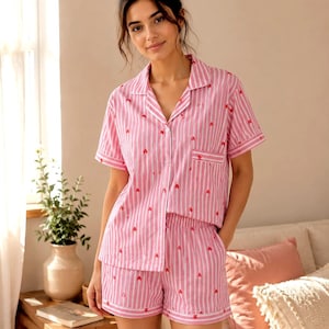 May include: A short-sleeved button-down pajama shirt and matching shorts set featuring a pink and white vertical striped pattern with small red heart icons scattered throughout the fabric. The shirt includes a notched collar and a single chest pocket.