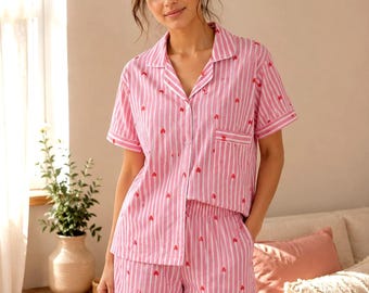 May include: A short-sleeved button-down pyjama shirt and matching shorts set featuring a pink and white vertical striped pattern with small red heart icons scattered throughout the fabric. The shirt includes a notched collar and a single chest pocket.