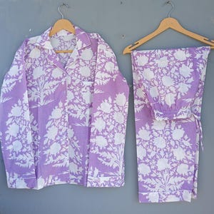 May include: A lavender and white floral pajama set. The set includes a button-down shirt with a chest pocket and matching trousers. The pajamas are made of a soft fabric and are displayed on wooden hangers against a grey background.