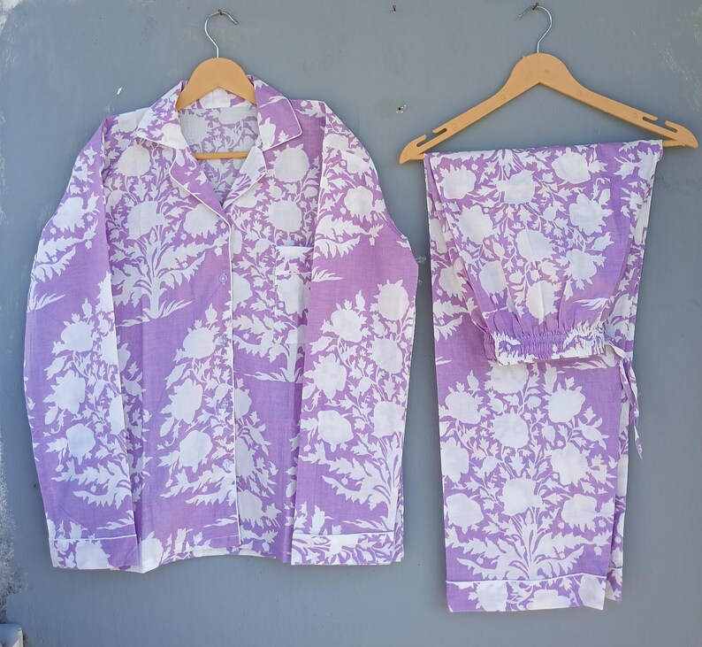 May include: A lavender and white floral pajama set. The set includes a button-down shirt with a chest pocket and matching trousers. The pajamas are made of a soft fabric and are displayed on wooden hangers against a grey background.