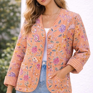 Puede incluir: Chaqueta con estampado floral melocotón con cuello en V, mangas tres cuartos y dos bolsillos delanteros. La chaqueta presenta un estampado floral colorido con flores naranjas, rosas, moradas y amarillas. Los puños y el ribete tienen rayas naranjas y blancas.
