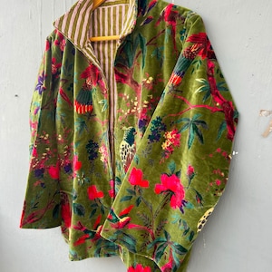 Op de afbeelding: Een jas met lange mouwen en een olijfgroene fluwelen buitenkant met een levendige print van kleurrijke vogels en bloemen. De binnenvoering is gemaakt van een wit en olijfgroen gestreepte stof, zichtbaar bij de kraag en de opening aan de voorzijde.
