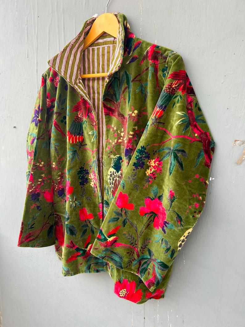 Op de afbeelding: Een jas met lange mouwen en een olijfgroene fluwelen buitenkant met een levendige print van kleurrijke vogels en bloemen. De binnenvoering is gemaakt van een wit en olijfgroen gestreepte stof, zichtbaar bij de kraag en de opening aan de voorzijde.