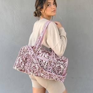 Peut inclure: Un sac de voyage matelassé arborant un motif floral imprimé en bloc de couleur bordeaux foncé et blanc. Le sac est doté de deux bandoulières robustes à rayures bordeaux et blanches, d'une forme rectangulaire et d'une fermeture éclair sur le dessus.
