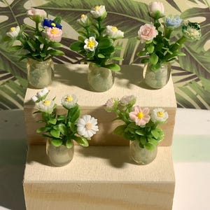 Peut inclure: Cinq arrangements floraux miniatures dans des vases en verre transparent, chacun doté de feuilles en plastique vert et de petites fleurs multicolores dans des tons de blanc, rose, bleu et jaune, présentés sur un support en bois clair à deux niveaux.