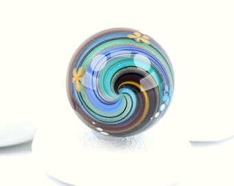 May include: A spherical glass marble featuring a swirling pattern of blue, teal, brown, and black colors. The surface is decorated with small, delicate yellow and light blue flower motifs and tiny white dots, with the text Madeline Bunyan visible.