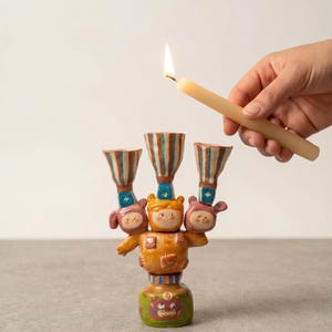 Puede incluir: Un candelabro de cerámica caprichoso con tres portavelas a rayas y una figura central. El candelabro está pintado en tonos naranja, azul y verde, con caras de dibujos animados. Se sostiene una vela encendida cerca.