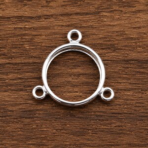 Könnte beinhalten: Ein silberfarbener Schmuckverbinder aus Metall mit einem zentralen kreisförmigen Rahmen und einem hohlen Innenraum. Das Bauteil verfügt über drei kleine, gleichmäßig verteilte kreisförmige Ösen, die vom Hauptring abstehen und zur Befestigung von Ketten dienen.