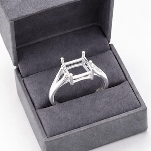 Könnte beinhalten: Ein silberner Ringrohling mit quadratischer Oberseite, präsentiert in einer grauen Ringschatulle. Der Ring hat ein geteiltes Schaftdesign und Krappen zur Aufnahme eines Edelsteins. Die Schatulle ist geöffnet und zeigt den Ring vor einem dunkelgrauen Innenraum.