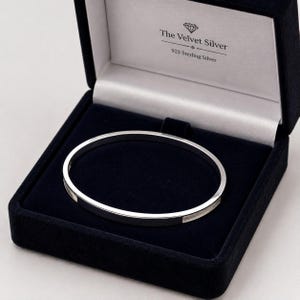 Könnte beinhalten: Ein polierter silberner Armreif liegt in einer dunkelblauen Schmuckschatulle aus Samt. Auf der Innenseite des Deckels steht der Text The Velvet Silver und 925 Sterling Silver mit einem Diamantsymbol. Der Armreif hat eine glatte, reflektierende Oberfläche.