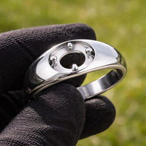 Könnte beinhalten: Ein polierter Silberring mit einem einzigartigen Design. Der Ring hat eine zentrale ovale Öffnung, umgeben von vier kleinen, erhabenen Stiften. Das Band ist glatt und abgerundet und reflektiert das Licht. Der Ring wird vor einem verschwommenen grünen Hintergrund gehalten.