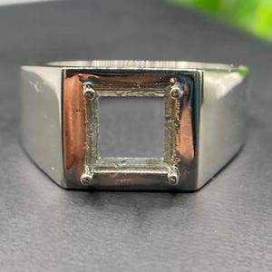 Könnte beinhalten: Ein polierter silberfarbener Metallring mit einer quadratischen Fassung und vier Krappen an den Ecken. Die Mitte der quadratischen Fassung ist leer und für die Aufnahme eines Edelsteins vorgesehen. Der Ring hat eine glatte Oberfläche.