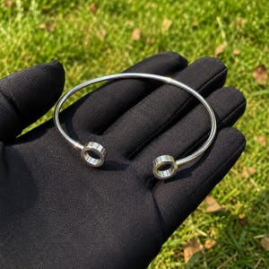 Könnte beinhalten: Ein Armreif aus silberfarbenem Metall mit einem glatten, dünnen Band, das an beiden Enden in einer kleinen, flachen, kreisförmigen Scheibe endet. Der Armreif hat ein offenes Design, wodurch er leicht um das Handgelenk gelegt werden kann.