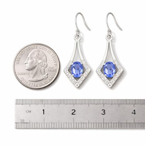 Könnte beinhalten: Silberne Ohrhänger mit einem blauen, ovalen Edelstein, umgeben von kleinen weißen Steinen, in einem rautenförmigen Rahmen. Die Ohrringe sind etwa 2.5 cm lang.