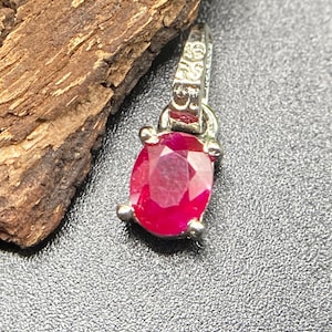 Könnte beinhalten: Ein silberner Anhänger mit einem oval geschliffenen, tief rosa-roten Edelstein, der von vier Krappen gehalten wird. Die Metallöse an der Oberseite ist mit der Zahl 925 geprägt, was auf Sterlingsilber hinweist. Das Metall ist poliert.