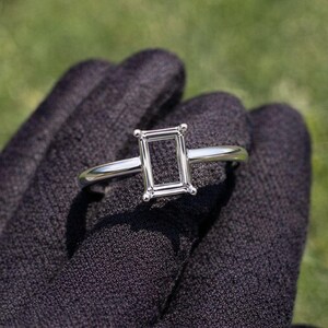 Könnte beinhalten: Ein silberner Ring mit einem Smaragdschliff. Der Ring wird vor einem dunklen Hintergrund gehalten, mit einem verschwommenen grünen Hintergrund. Die Fassung des Rings ist rechteckig und das Band ist dünn und poliert.