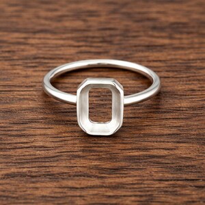 Könnte beinhalten: Ein silberner Ring mit rechteckiger Lünettenfassung. Der Ring hat ein schmales, abgerundetes Band, das zur offenen rechteckigen Fassung führt. Der Ring wird auf einer Holzoberfläche gezeigt.