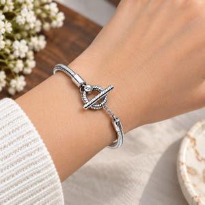 Könnte beinhalten: Ein silbernes Schlangenkettarmband mit einem Knebelverschluss. Der kreisförmige Knebelring ist mit der Zahl 925 graviert, was auf eine Zusammensetzung aus Sterlingsilber hinweist. Das Armband hat eine texturierte, seilartige Optik.