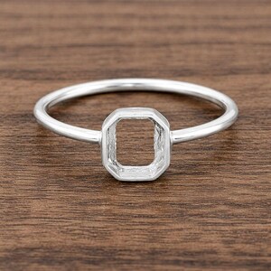May include: A silver ring with a geometric, octagonal setting. The ring band is thin and circular, leading to the open, square-shaped setting. The ring is made of a shiny, metallic material.