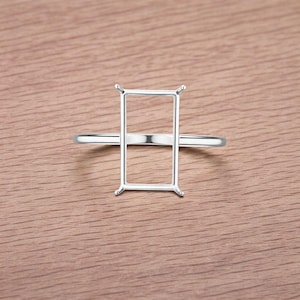 Könnte beinhalten: Ein silberner Ring mit rechteckiger Fassung, zur Aufnahme eines Edelsteins. Die Fassung hat vier Krappen an jeder Ecke, um den Stein zu sichern. Der Ring ist dünn und glatt, präsentiert auf einer Holzoberfläche.