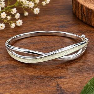 Könnte beinhalten: Ein silberfarbener Klapparmreif mit polierter Metalloberfläche und einem zentralen, überkreuzten Designelement mit einer texturierten, blassgelben Einlage. Das Armband verfügt über einen kleinen kugelförmigen Verschlussmechanismus an einer Seite.