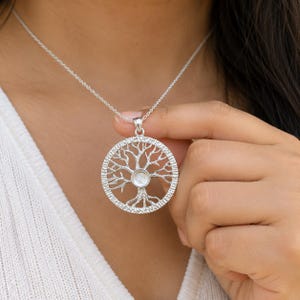 Könnte beinhalten: Eine silberfarbene, kreisförmige Anhängerkette mit einem komplexen Lebensbaum-Design und einer zentralen Steinfassung. Der Anhänger hängt an einer feinen silberfarbenen Kette und weist ein strukturiertes Metallfinish am äußeren Ring auf.