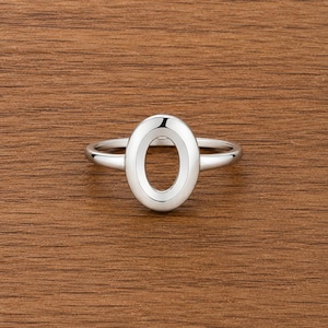 Könnte beinhalten: Ein silberner Ring mit dem Buchstaben "O" in Großbuchstaben. Der Ring hat eine polierte Oberfläche und ein schlichtes Band. Der Ring wird auf einer Holzoberfläche präsentiert.