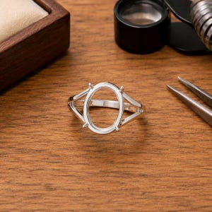 Könnte beinhalten: Eine silberne Ringfassung mit einem ovalen Mittelteil und vier Krappen. Der Ring hat ein geteiltes Schaftdesign und wird auf einer Holzoberfläche mit Schmuckwerkzeugen im Hintergrund präsentiert.