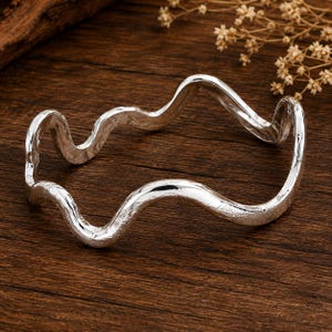 May include: A silver-coloured bangle bracelet featuring a continuous, undulating wave pattern. The metal surface has a textured, slightly matte finish throughout, creating a consistent appearance along the entire length of the circular jewellery piece.