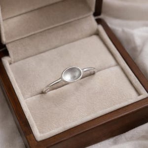 Könnte beinhalten: Ein silberner Ring mit ovaler Fassung, präsentiert in einer Holzschatulle mit beigem Innenraum. Der Ring hat ein schmales Band, das zu einem prominenten ovalen Mittelstück führt. Der Ring ist schlicht und elegant gestaltet.