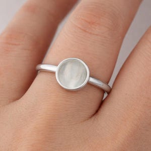Könnte beinhalten: Ein silberner Ring mit einer kreisförmigen, flachen Fassung. Der Ring ist schlicht und elegant, mit einem dünnen Band und einer glatten, polierten Oberfläche. Die Fassung ist für einen Stein oder eine andere Verzierung gedacht.