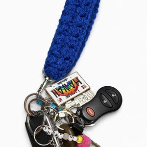 Puede incluir: Un llavero de pulsera tejido en crochet azul con una argolla metálica y varios accesorios, incluyendo un dije de placa de matrícula de Hawái, una llave de coche negra, un pequeño control remoto negro y cuentas que forman la palabra TEADU.