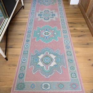 Könnte beinhalten: Ein rechteckiger, rosa Läufer mit einem geometrischen Muster in Blau-, Türkis- und Weißtönen. Der Teppich hat ein zentrales Design mit sich wiederholenden Motiven und einen Rand mit kreisförmigen und rautenförmigen Formen. Er liegt auf einem Holzboden.