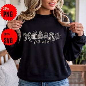Peut inclure: Un sweat-shirt noir à manches longues avec une impression graphique blanche représentant un nœud, une citrouille, un bonnet en tricot, une tasse de café, une écharpe rayée et une feuille d'érable, avec le texte "fall vibes" en cursive.