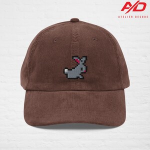 K&ouml;nnte beinhalten: Braune Cord-Baseballkappe mit einem gestickten, pixeligen grauen Hasen mit rosa Akzenten. Die Kappe hat einen gebogenen Schirm und einen Knopf oben. Das Logo "A/D ATELIER DECODE" ist sichtbar.