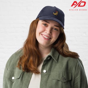 K&ouml;nnte beinhalten: Marineblaue Cord-Baseballkappe mit einem kleinen gestickten Design eines Cartoon-Tiers mit Hut. Die Kappe wird von einer Person getragen, die eine gr&uuml;ne Cordjacke tr&auml;gt. Das Atelier Decode-Logo ist sichtbar.
