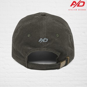 K&ouml;nnte beinhalten: Olivgr&uuml;ne Cord-Baseballkappe mit gebogenem Schirm und verstellbarem Riemen. Die Kappe hat die gestickten Initialen "A/D" auf der Vorder- und R&uuml;ckseite. Der Markenname "ATELIER DECODE" ist oben rechts aufgedruckt.
