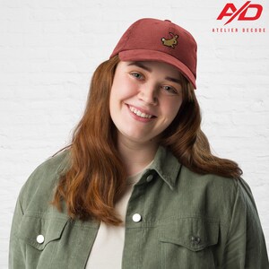 K&ouml;nnte beinhalten: Eine rotbraune Cord-Baseballkappe mit einem gestickten Tierdesign. Die Kappe wird von einer Person getragen, die eine gr&uuml;ne Cordjacke tr&auml;gt. Das Atelier Decode-Logo ist oben rechts sichtbar. Die Kappe ist ein stilvolles Accessoire.