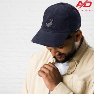 K&ouml;nnte beinhalten: Marineblaue Cord-Baseballkappe mit einem pixeligen grauen Hasen-Motiv auf der Vorderseite. Die Kappe hat einen gebogenen Schirm und eine strukturierte Krone. Das Logo "A/D ATELIER DECODE" ist oben rechts aufgedruckt.