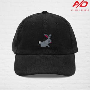 K&ouml;nnte beinhalten: Schwarze Cord-Baseballkappe mit einem gestickten pixeligen grauen Hasen mit rosa Akzenten. Die Kappe hat einen gebogenen Schirm und einen Knopf oben. Das rote Logo lautet "A/D ATELIER DECODE".