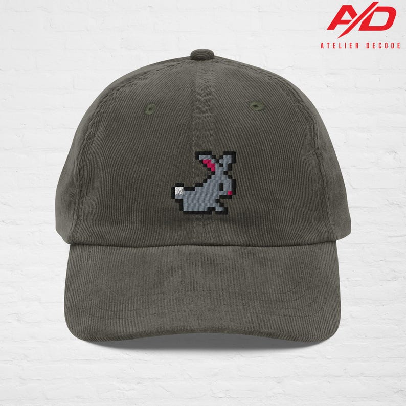 K&ouml;nnte beinhalten: Graue Cord-Baseballkappe mit einem pixeligen grauen Hasen mit rosa Akzenten. Die Kappe hat einen gebogenen Schirm und einen Knopf oben. Das Logo "A/D ATELIER DECODE" ist oben rechts aufgedruckt.