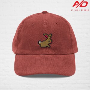 K&ouml;nnte beinhalten: Eine ziegelrote Cord-Baseballkappe mit einer pixeligen, braunen Hasenstickerei. Die Kappe hat einen gebogenen Schirm und einen Knopf oben. Das Logo "A/D ATELIER DECODE" ist oben rechts aufgedruckt.