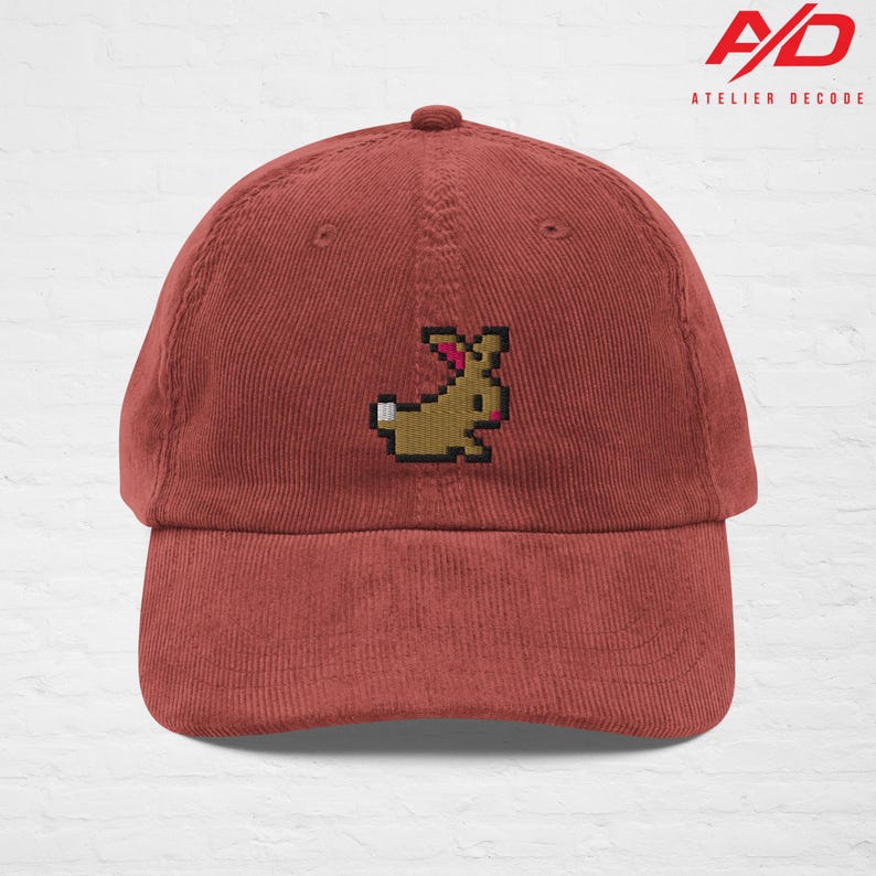 K&ouml;nnte beinhalten: Eine ziegelrote Cord-Baseballkappe mit einer pixeligen, braunen Hasenstickerei. Die Kappe hat einen gebogenen Schirm und einen Knopf oben. Das Logo "A/D ATELIER DECODE" ist oben rechts aufgedruckt.