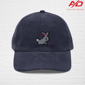 K&ouml;nnte beinhalten: Marineblaue Cord-Baseballkappe mit einem gestickten pixeligen grauen Hasen mit rosa Akzenten. Die Kappe hat einen gebogenen Schirm und einen Knopf oben. Das Atelier Decode-Logo ist sichtbar.