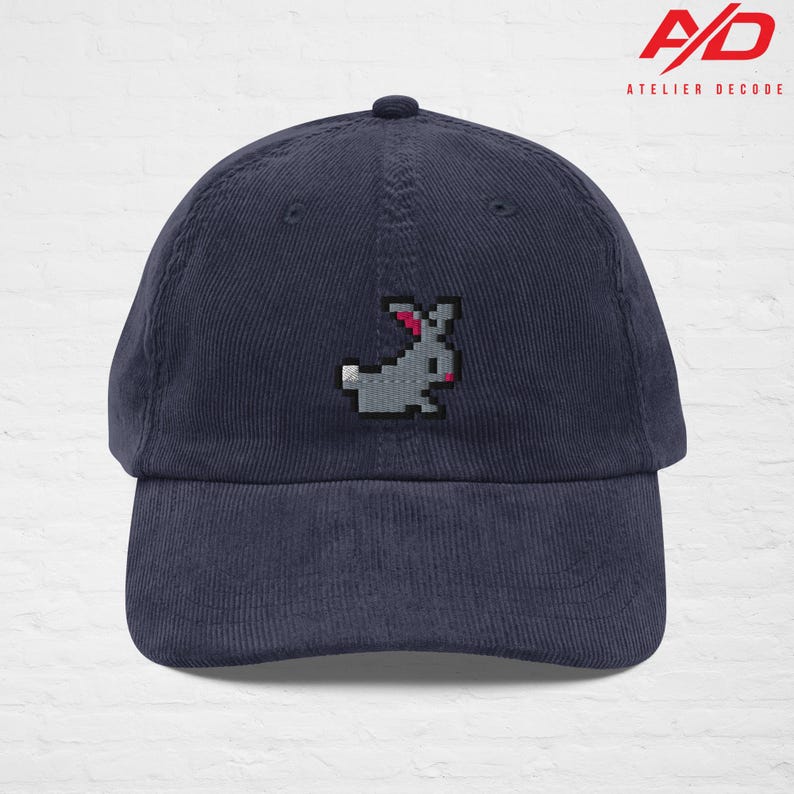 K&ouml;nnte beinhalten: Marineblaue Cord-Baseballkappe mit einem gestickten pixeligen grauen Hasen mit rosa Akzenten. Die Kappe hat einen gebogenen Schirm und einen Knopf oben. Das Atelier Decode-Logo ist sichtbar.
