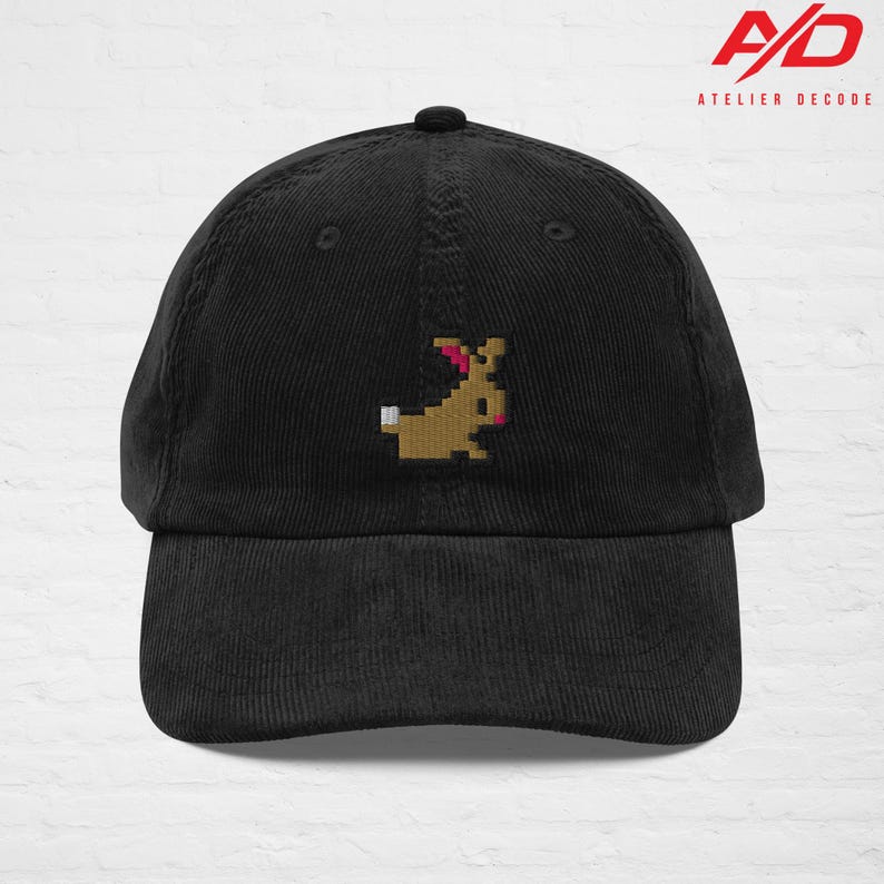 K&ouml;nnte beinhalten: Schwarze Cord-Baseballkappe mit einem gestickten pixeligen Hasen-Design in Braun, Rosa und Wei&szlig;. Die Kappe hat einen gebogenen Schirm und einen Knopf oben. Das Markenlogo "A/D ATELIER DECODE" ist oben rechts aufgedruckt.