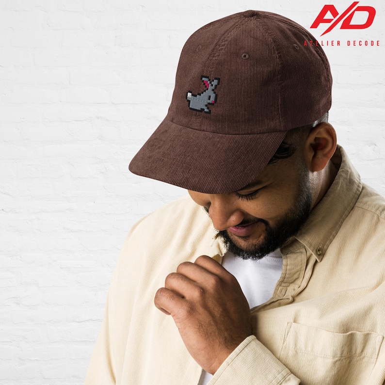 K&ouml;nnte beinhalten: Braune Cord-Baseballkappe mit einem pixeligen grauen Hasen mit rosa Schleife. Die Kappe hat einen gebogenen Schirm und eine strukturierte Krone. Das Logo "A/D Atelier Decode" ist oben rechts aufgedruckt.