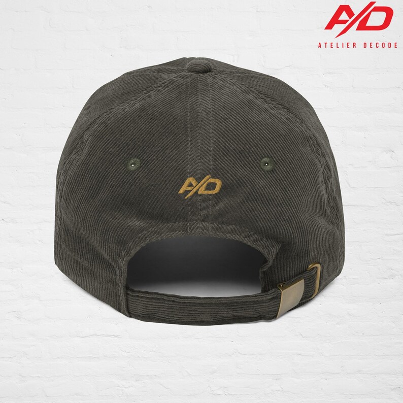 K&ouml;nnte beinhalten: Dunkel olivgr&uuml;ne Cord-Baseballkappe mit gebogenem Schirm und verstellbarem Riemen. Auf der R&uuml;ckseite der Kappe befindet sich ein goldfarbenes, gesticktes "A/D"-Logo. Der Markenname "ATELIER DECODE" ist in Rot und Wei&szlig; aufgedruckt.