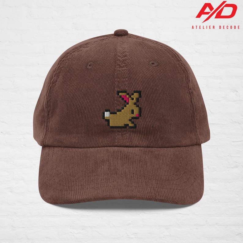 K&ouml;nnte beinhalten: Braune Cord-Baseballkappe mit einem gestickten pixeligen Hasen-Design. Die Kappe hat einen gebogenen Schirm und einen Knopf oben. Das Logo "A/D ATELIER DECODE" ist oben rechts aufgedruckt.