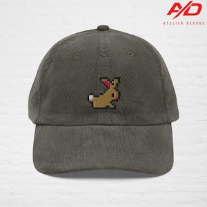 K&ouml;nnte beinhalten: Olivgr&uuml;ne Cord-Baseballkappe mit einem gestickten, pixeligen Hasen-Design in Braun, Wei&szlig; und Rosa. Der Markenname "ATELIER DECODE" ist rechts oben in Rot und Wei&szlig; aufgedruckt.